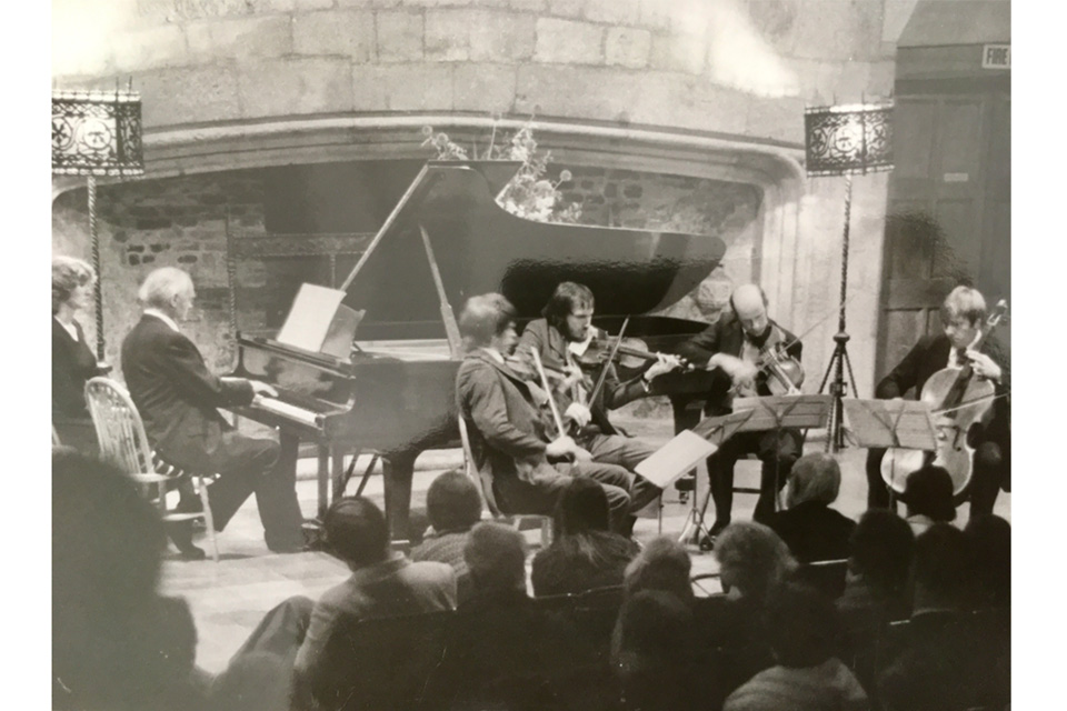 An old photograph of a group of male musicians, wearing smart suits, performing on string instruments and a piano, in front of an audience.